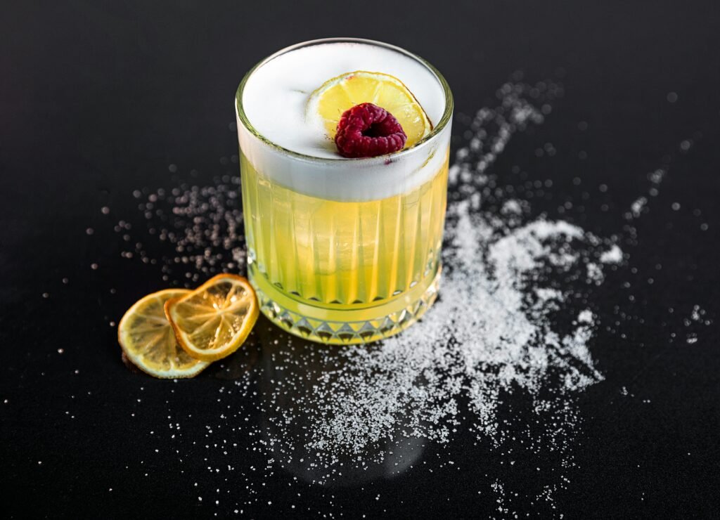 A vibrant lemon cocktail garnished with raspberry, set against a black background with sugar detailing.