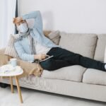 Sick man with mask and scarf resting on couch at home, feeling unwell.