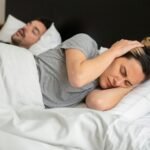 A woman looks frustrated in bed as her partner snores loudly, highlighting sleep disturbance issues.