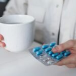 Close-up of a woman holding a hot drink and blister pack of blue capsules indoors, suggesting illness or treatment.