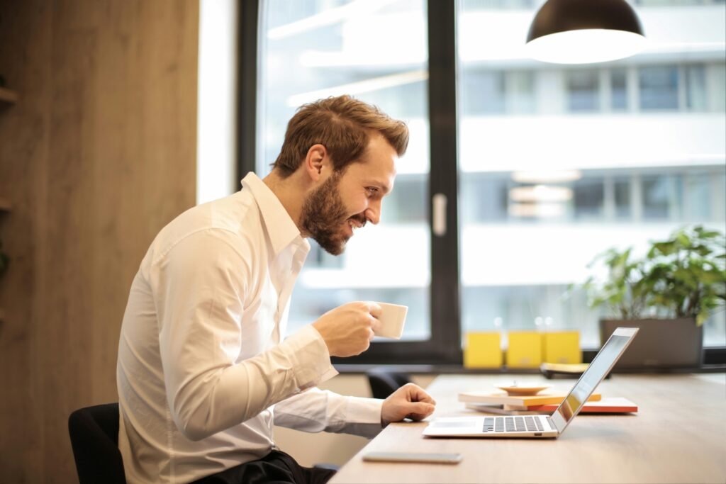 Man in office with coffee, smiling while working at laptop, captures the essence of remote work.